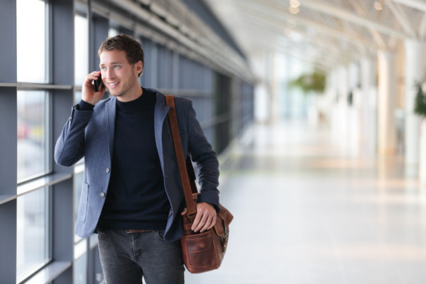 Urban business man talking on smart phone traveling walking inside in airport. Casual young businessman wearing suit jacket and shoulder bag. Handsome male model in his 20s.